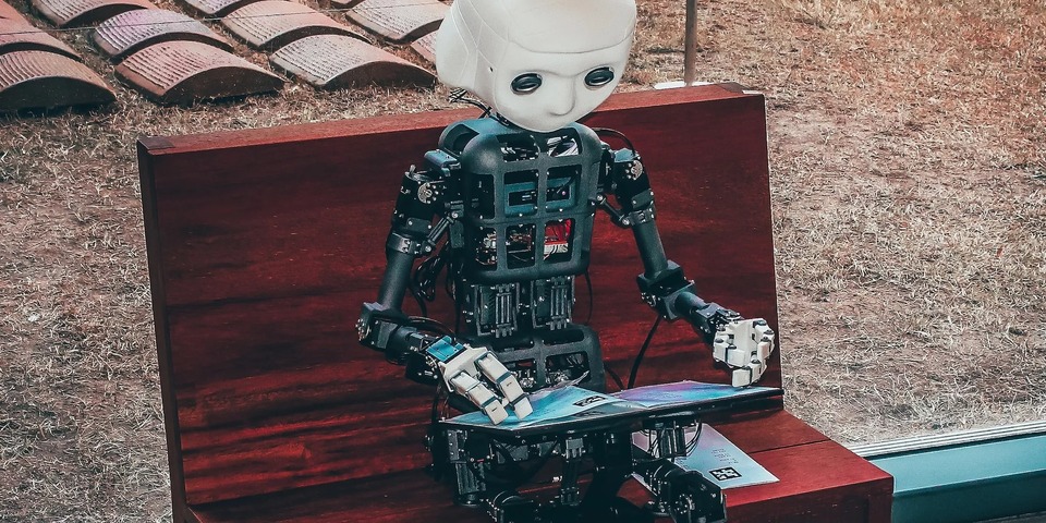 A humanoid robot sits on a wooden bench reading a magazine.